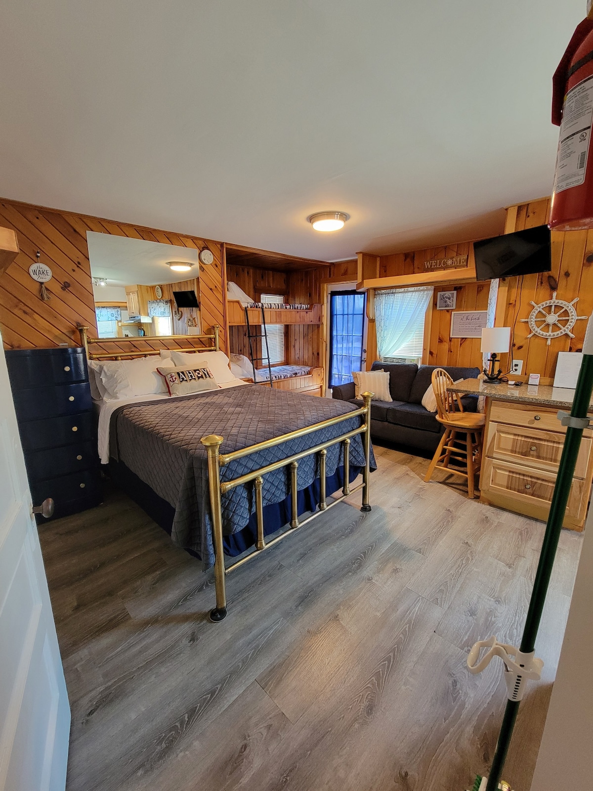 A studio space features a full size bed with a dark blue quilt and decorative pillows, complemented by wood paneling. A cozy seating area is present with a chair and a small table. Natural light enters through a window with sheer curtains.