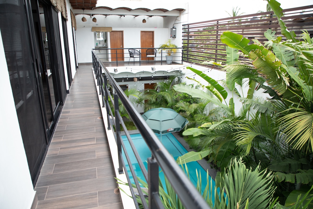 A balcony walkway is seen leading to rooms, flanked by lush green foliage. The view overlooks a private swimming pool surrounded by tropical plants. Natural light spills into the area, enhancing the serene atmosphere of the space.
