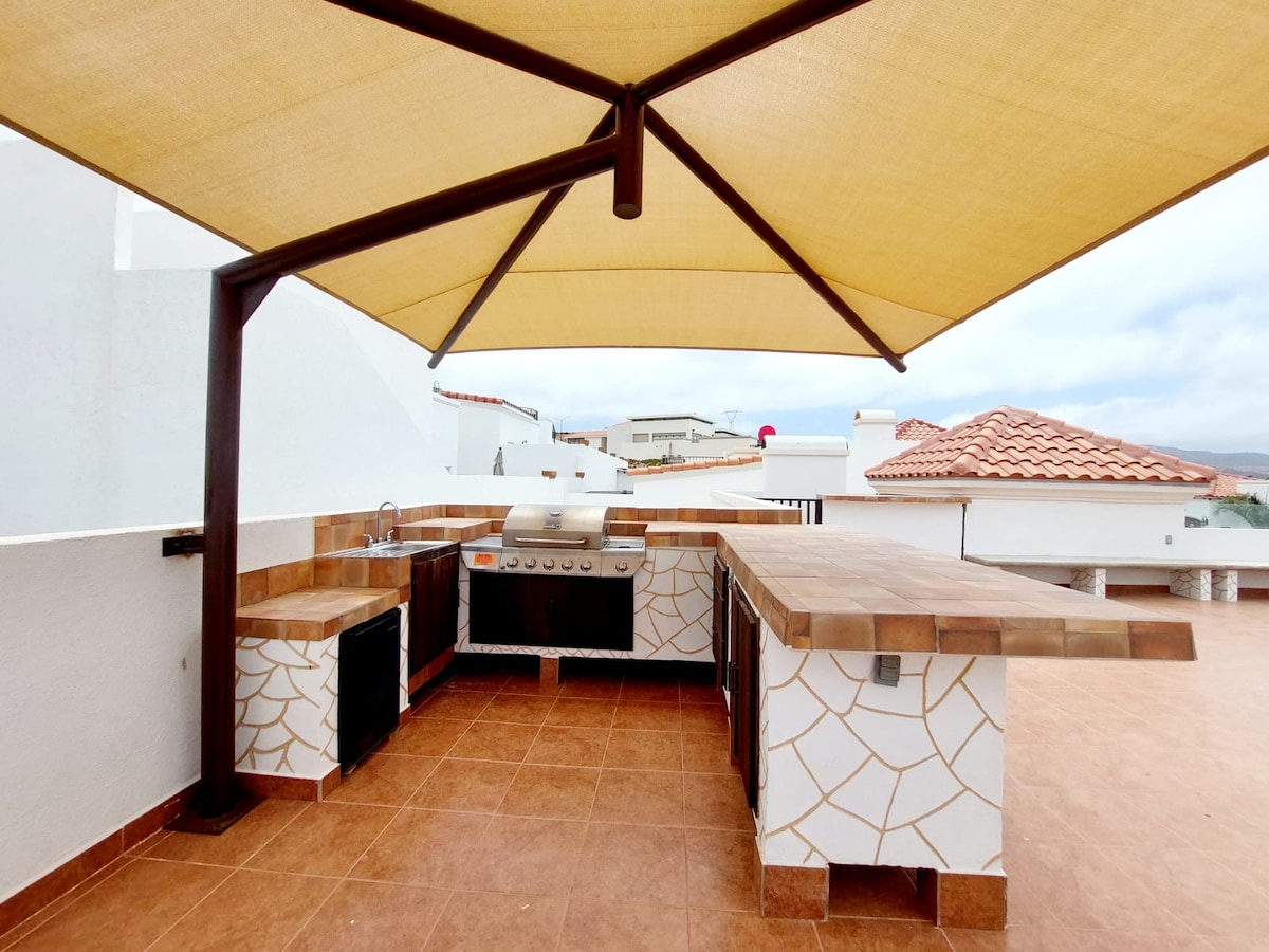 A rooftop grilling area features a covered space with a large aluminum grill and ample counter space. The tan tiles and patterned backsplash complement the surrounding walls, creating an open and functional environment for outdoor cooking.