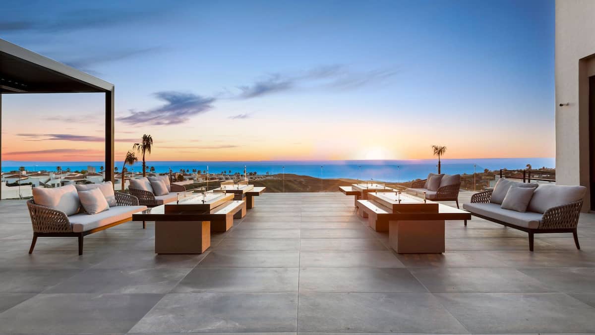 A spacious terrace is furnished with multiple seating arrangements, featuring comfortable sofas and coffee tables. The ocean view stretches to the horizon, where a vibrant sunset casts warm hues across the sky. The setting offers an inviting space for relaxation.