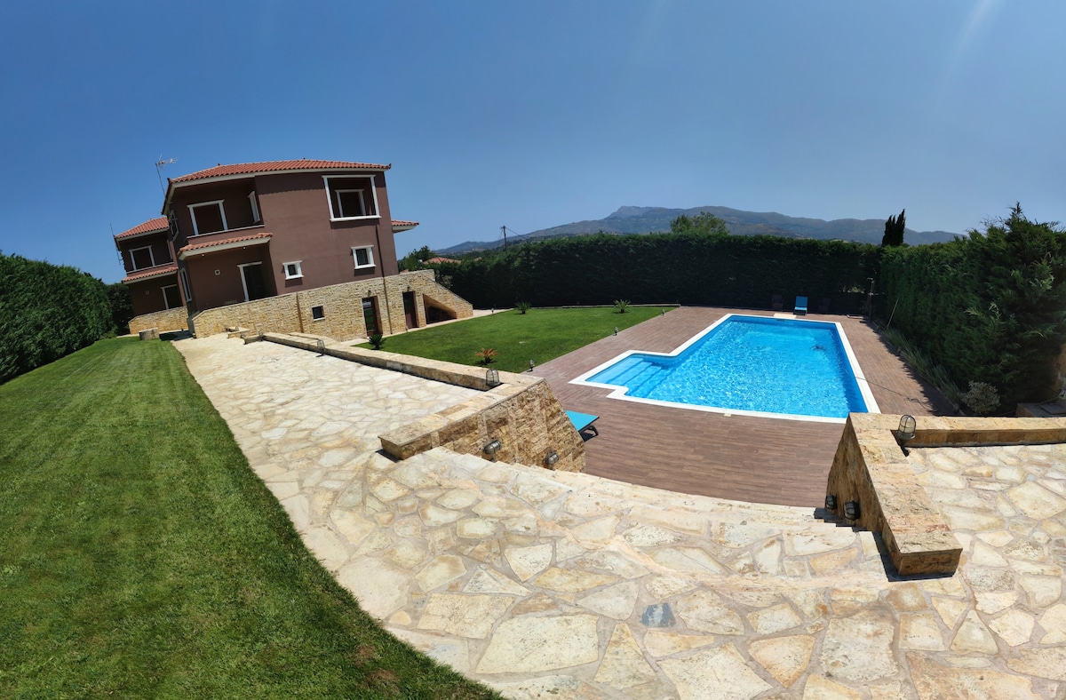 A spacious outdoor area features a crystal-clear swimming pool bordered by a stone deck. Lush green grass surrounds the pool, and a two-story villa is positioned nearby, framed by tall hedges and scenic views of distant mountains.