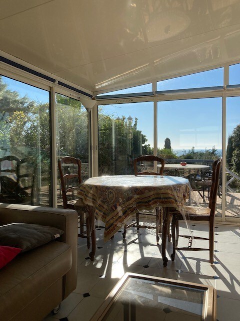 A sunlit dining area is surrounded by large glass windows, offering views of the garden and ocean. A round table is set with intricate table linens, complemented by wooden chairs. Natural light illuminates the space, enhancing the serene atmosphere.