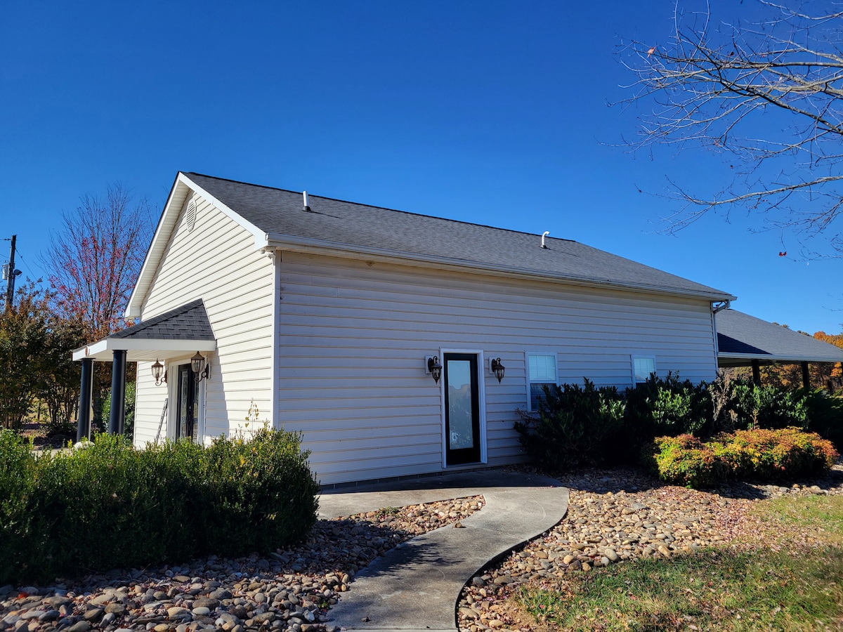 The exterior of the guesthouse is showcased, featuring a light-colored vinyl siding and a charming covered entrance. Surrounding landscaping includes neatly trimmed shrubs and a pebbled pathway, leading to the entrance. A clear blue sky completes the serene countryside setting.