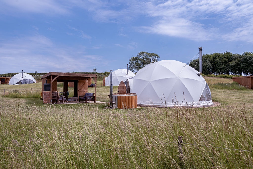 Two white domes stand amidst tall grass in a spacious outdoor setting. A wooden gazebo with seating is visible beside a hot tub, providing a serene space for relaxation. The backdrop features trees and a clear blue sky.