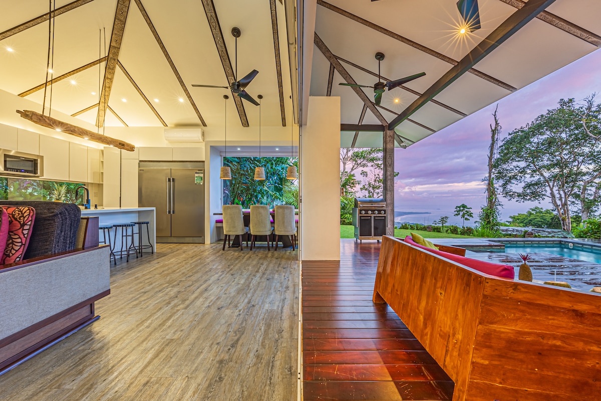 An open-concept living area features a modern kitchen with a large island and sleek appliances. The space transitions seamlessly to an outdoor deck, enhanced by a plunge pool and a gas grill, with views of the surrounding nature and coastline.
