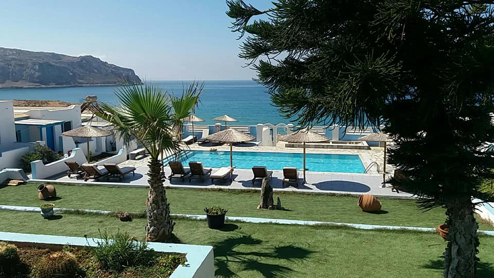 A tranquil outdoor area is depicted with a swimming pool surrounded by sun loungers under thatched umbrellas. Lush green lawns and palm trees are visible, along with a backdrop of clear blue waters and a rocky coastline.