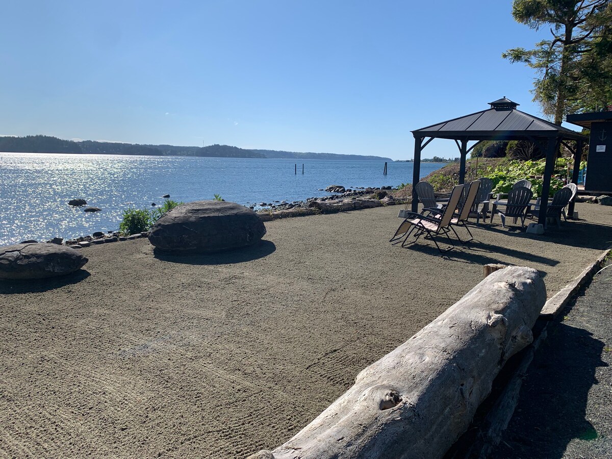 A serene waterfront space features a gazebo and several comfortable chairs arranged on a smooth surface. Large rocks and driftwood are positioned around the area, overlooking a sparkling, calm body of water with trees framing the background.