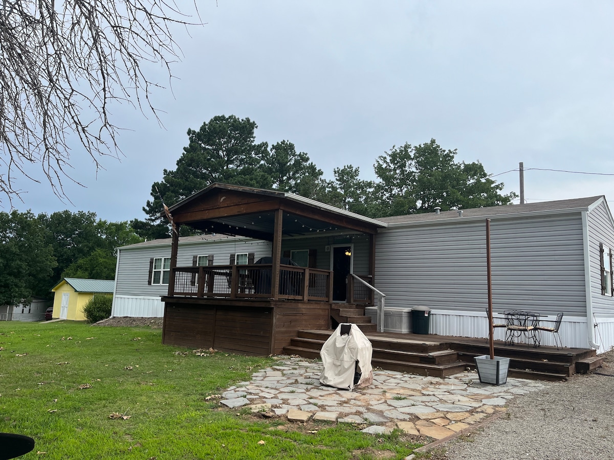 A modern mobile home is viewed from the exterior, featuring a wooden deck with steps leading to the entrance. Soft outdoor lighting adorns the deck area, and a stone patio is visible in front, surrounded by grass. Additional structures can be seen in the background.