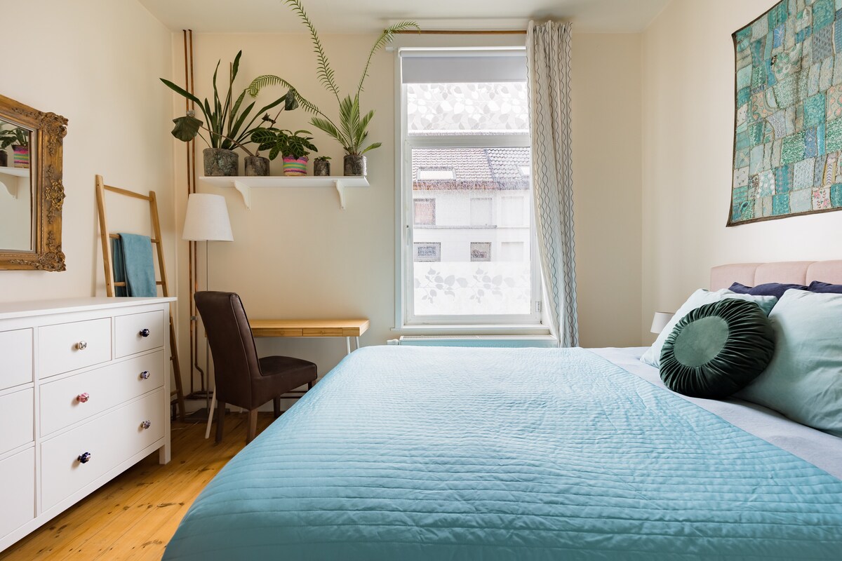 A softly lit bedroom features a spacious bed covered in a light blue quilt. A decorative pillow adds texture. A mirrored dresser and a desk chair complement a wooden desk, while a plant display adorns the shelf by the window, allowing natural light to filter in.