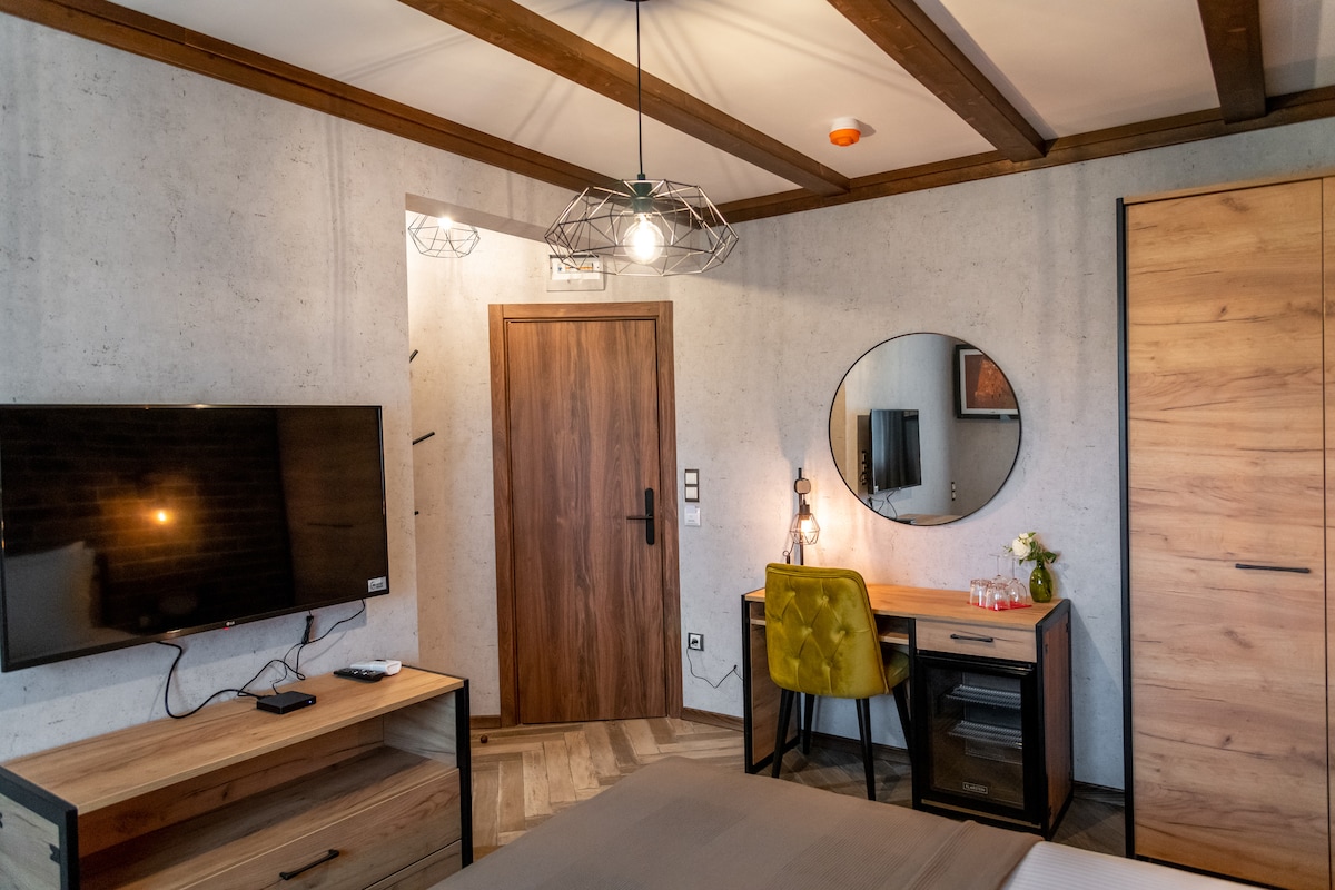A well-appointed room features a comfortable double bed and a wooden desk paired with an ergonomic chair. A flat-screen television is mounted on the wall across from the bed, while a round mirror and small decorative elements complete the inviting atmosphere.