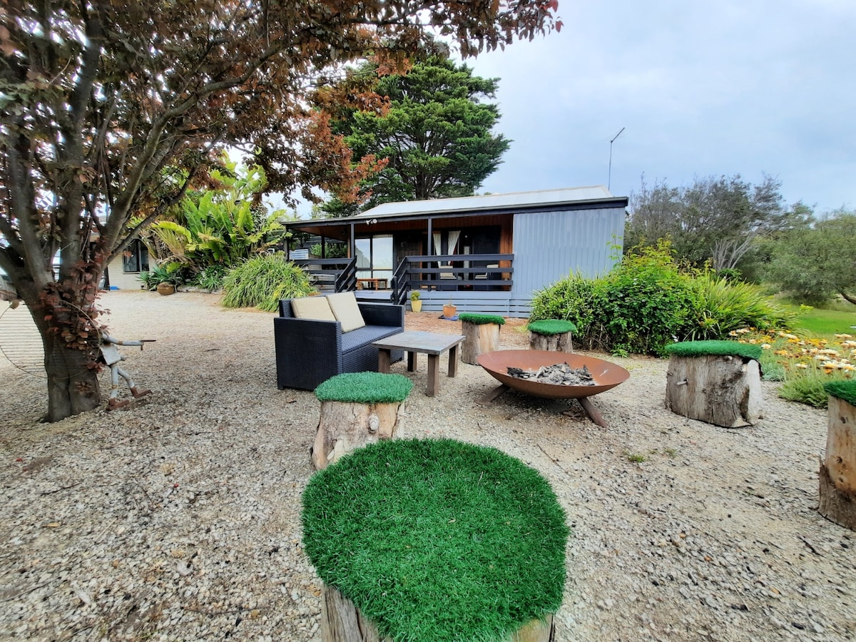 An outdoor seating area features a dark wicker sofa paired with circular green grass-like stools. A fire pit is positioned on a gravel surface, surrounded by lush greenery and trees. The cabin is visible in the background, set against a cloudy sky.