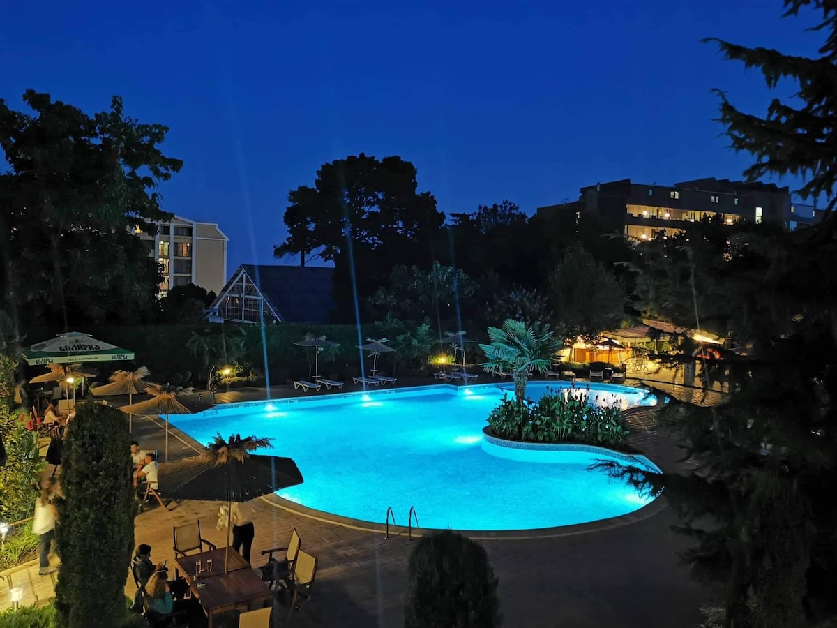 The outdoor swimming pool is illuminated in vibrant blue light, surrounded by lush greenery and palm trees. Comfortable lounge chairs are arranged along the pool's edge, while tables under umbrellas create a relaxed dining area nearby, enhancing the serene evening atmosphere.