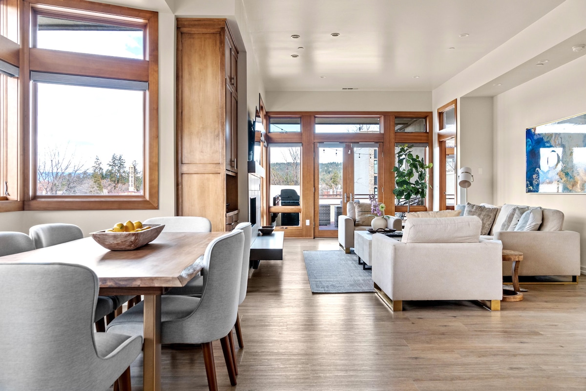 The living area features large windows allowing natural light to fill the space, revealing a comfortable seating arrangement with a sectional sofa and stylish armchairs. A dining table with upholstered chairs sits adjacent, while a modern fireplace adds warmth and atmosphere.