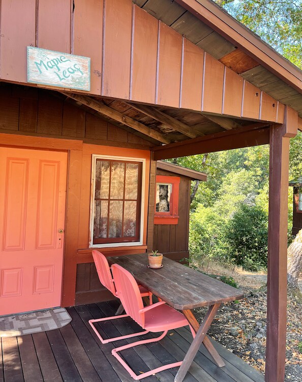 The front porch of the Maple Leaf Cabin features a rustic wooden table accompanied by two pink chairs. A small potted plant rests on the table. The cabin's door is painted in a bright coral color, and a sign labeled 'Maple Leaf' hangs above.
