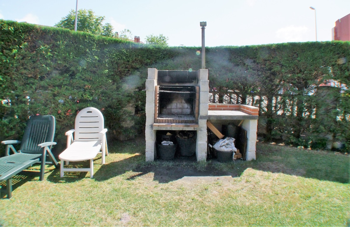 An outdoor barbecue area is visible, featuring a stone grill setup with a wooden prep table beside it. Two lounge chairs are positioned nearby on the grassy lawn, surrounded by a well-maintained hedge for privacy.