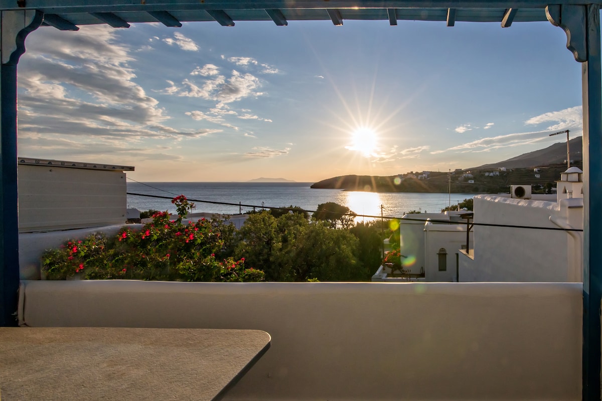 A panoramic view captures the sun setting over the sea, casting a warm glow across the water. Vibrant flowers add color in the foreground, while hills and buildings outline the coastal landscape in the distance.