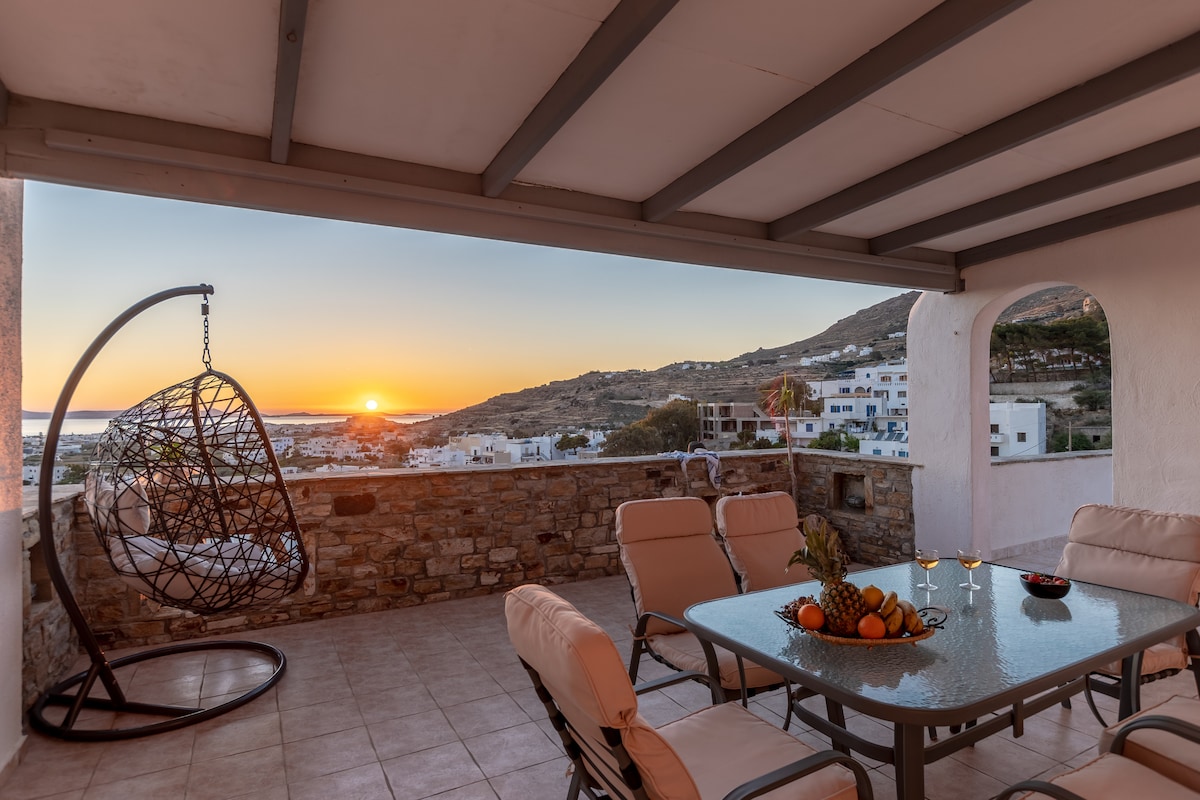 A spacious outdoor terrace features a glass table surrounded by comfortable chairs. A hanging chair is positioned nearby, creating a relaxed seating area. The view of the sunset over the Aegean Sea can be seen in the background, enhancing the peaceful atmosphere.