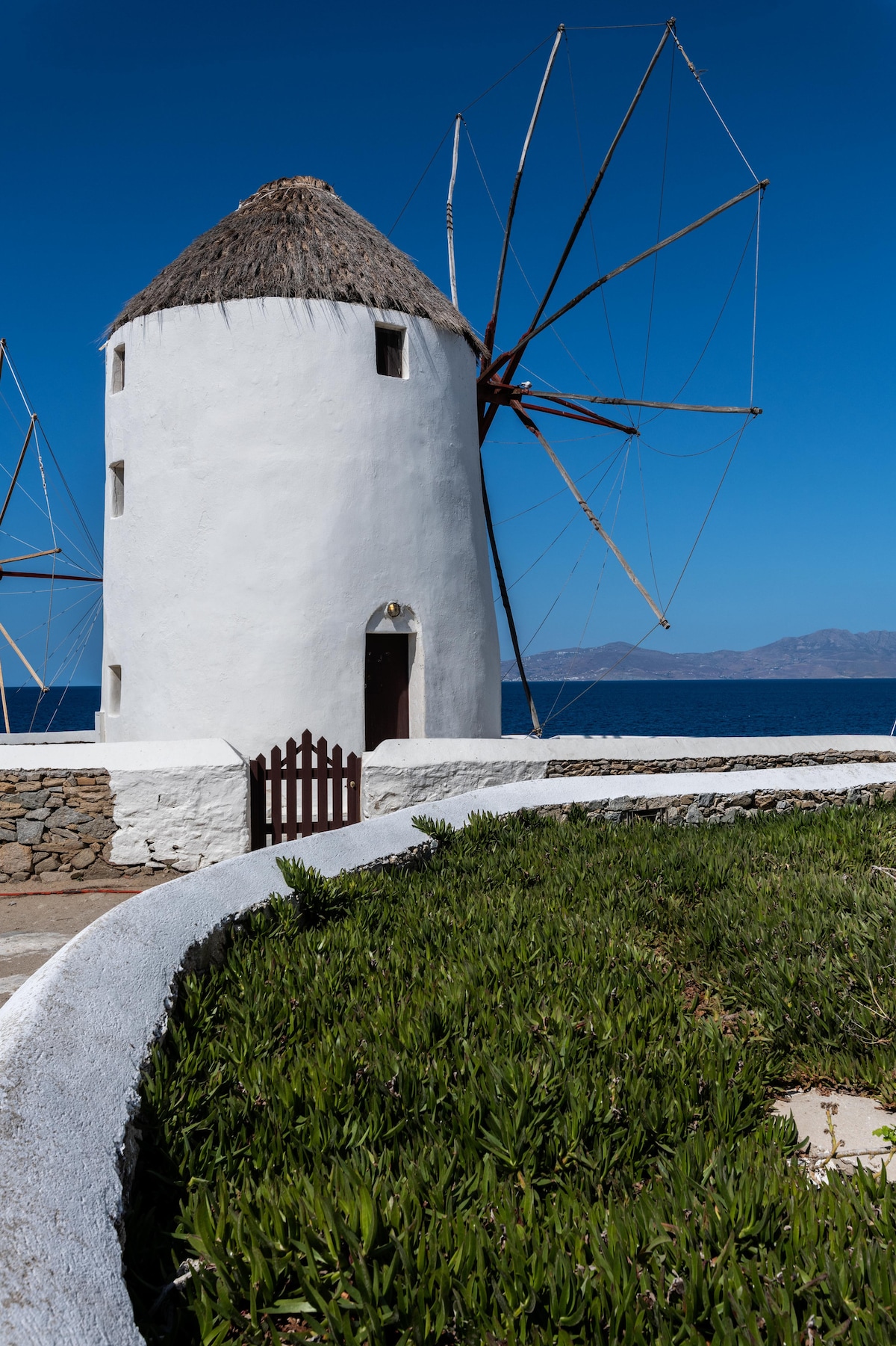 The image features the Iconic Windmill, showcasing its traditional whitewashed structure and thatched roof. A wooden door is visible at the base, alongside a neatly maintained walled garden with lush greenery. The backdrop includes a vibrant blue sky and the tranquil Aegean Sea.