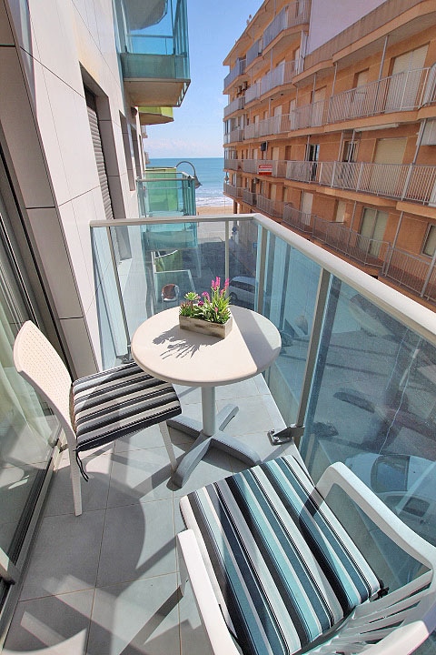 A small balcony is visible, featuring a round table with a potted plant and two striped chairs. The ocean can be seen in the background, along with a sandy beach and buildings lining the street below.