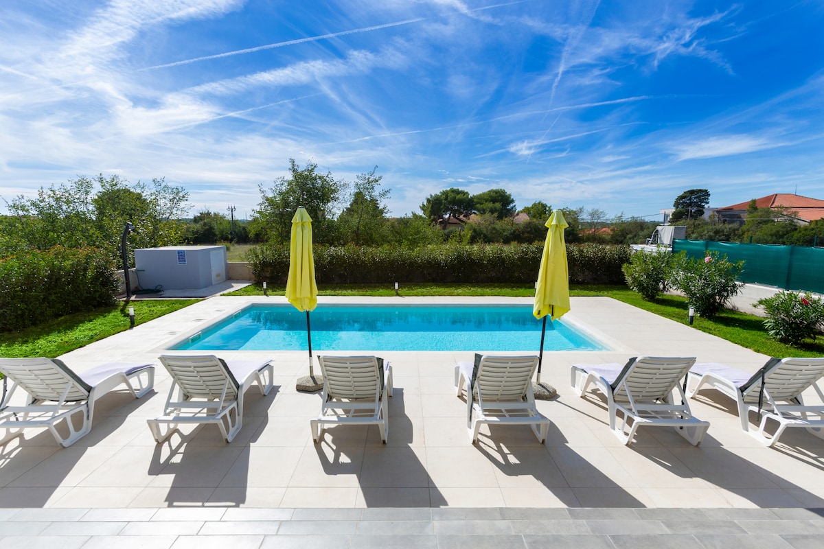 An inviting pool area is surrounded by a spacious patio, featuring multiple lounge chairs and two yellow umbrellas providing shade. Lush greenery borders the area, with the sky displaying light clouds and blue tones, creating a serene outdoor setting.