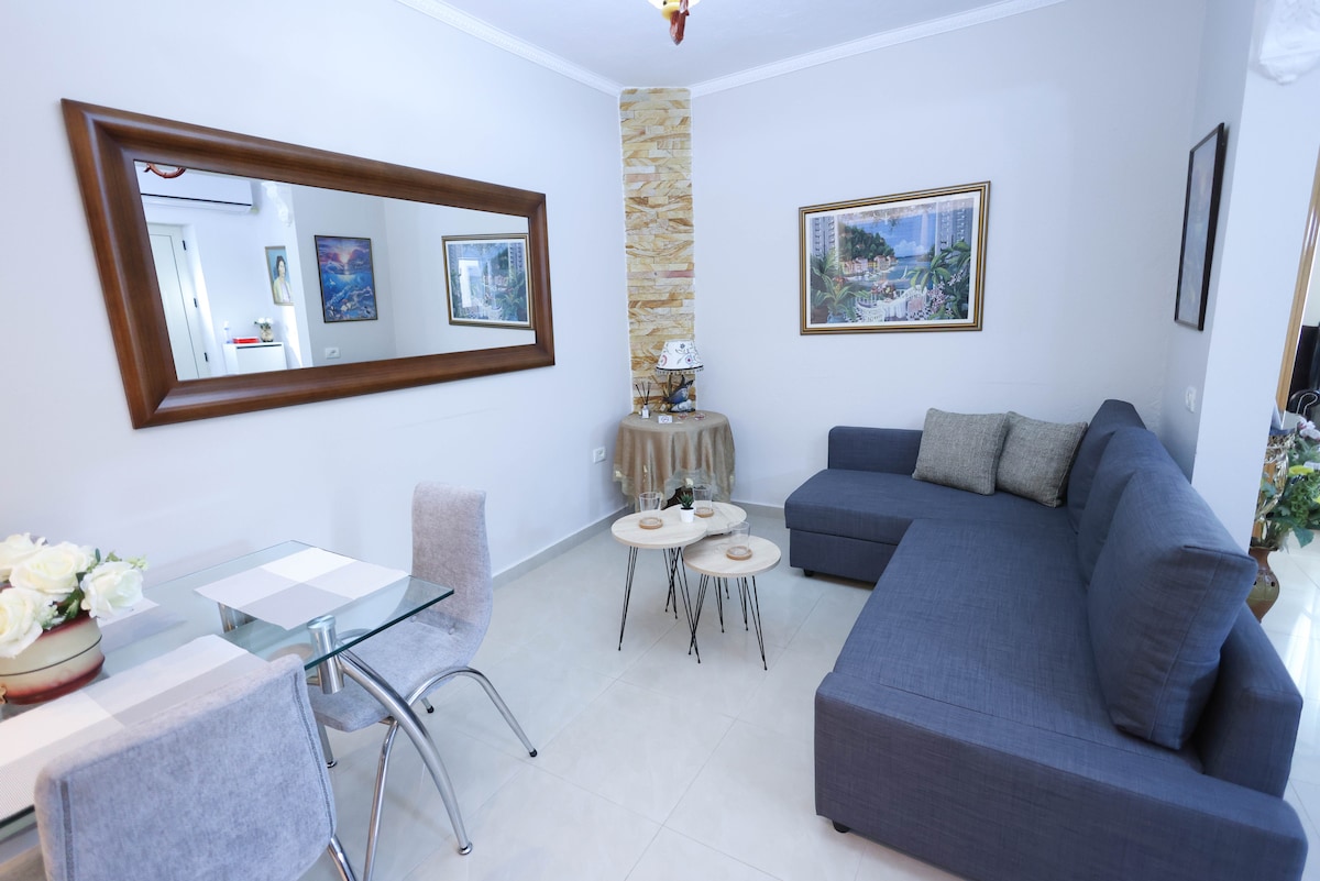 A cozy living area features a dark blue sectional sofa with decorative cushions. A round table is set with light-colored tableware, and two small coffee tables are positioned nearby. A large mirror enhances the space, reflecting the light from the surrounding area.