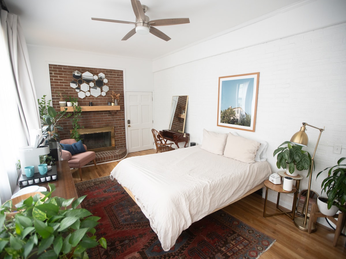 A mid century modern bedroom features a spacious bed adorned with neutral linens. Natural light illuminates the room, highlighting a brick fireplace and decorative wall mirrors. A comfortable seating area with a chair and table, along with lush greenery, enhances the serene atmosphere.