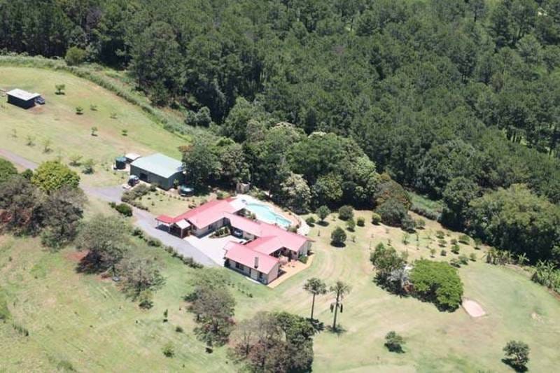 An aerial view captures a sprawling property set within lush greenery. The main building is surrounded by landscaped gardens, featuring a swimming pool and a variety of trees. Additional structures are visible, along with a meandering driveway that integrates into the serene landscape.