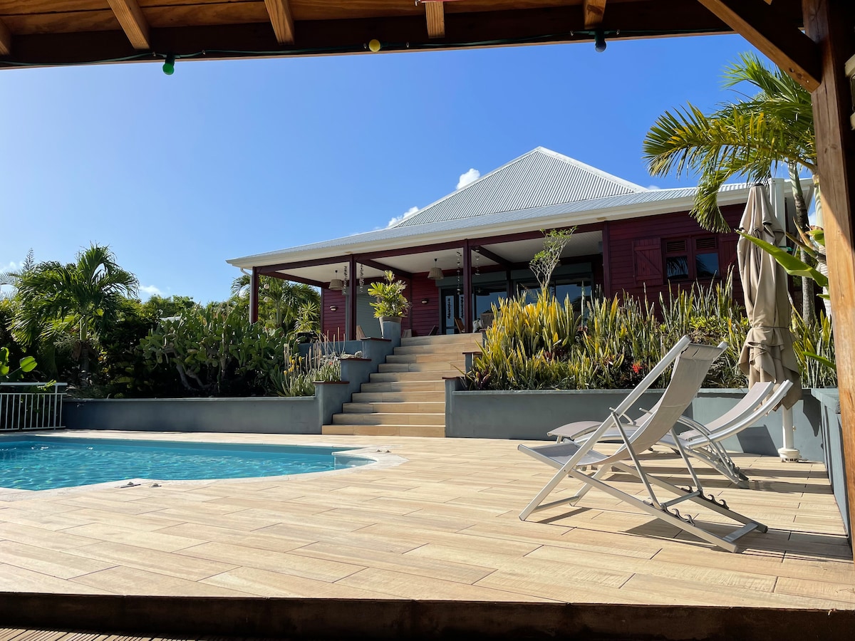 A vibrant exterior of a house is showcased, featuring a spacious deck and a swimming pool. Steps lead up from the pool area to the house, surrounded by tropical greenery. Lounge chairs are positioned beside the pool, inviting relaxation under a clear blue sky.