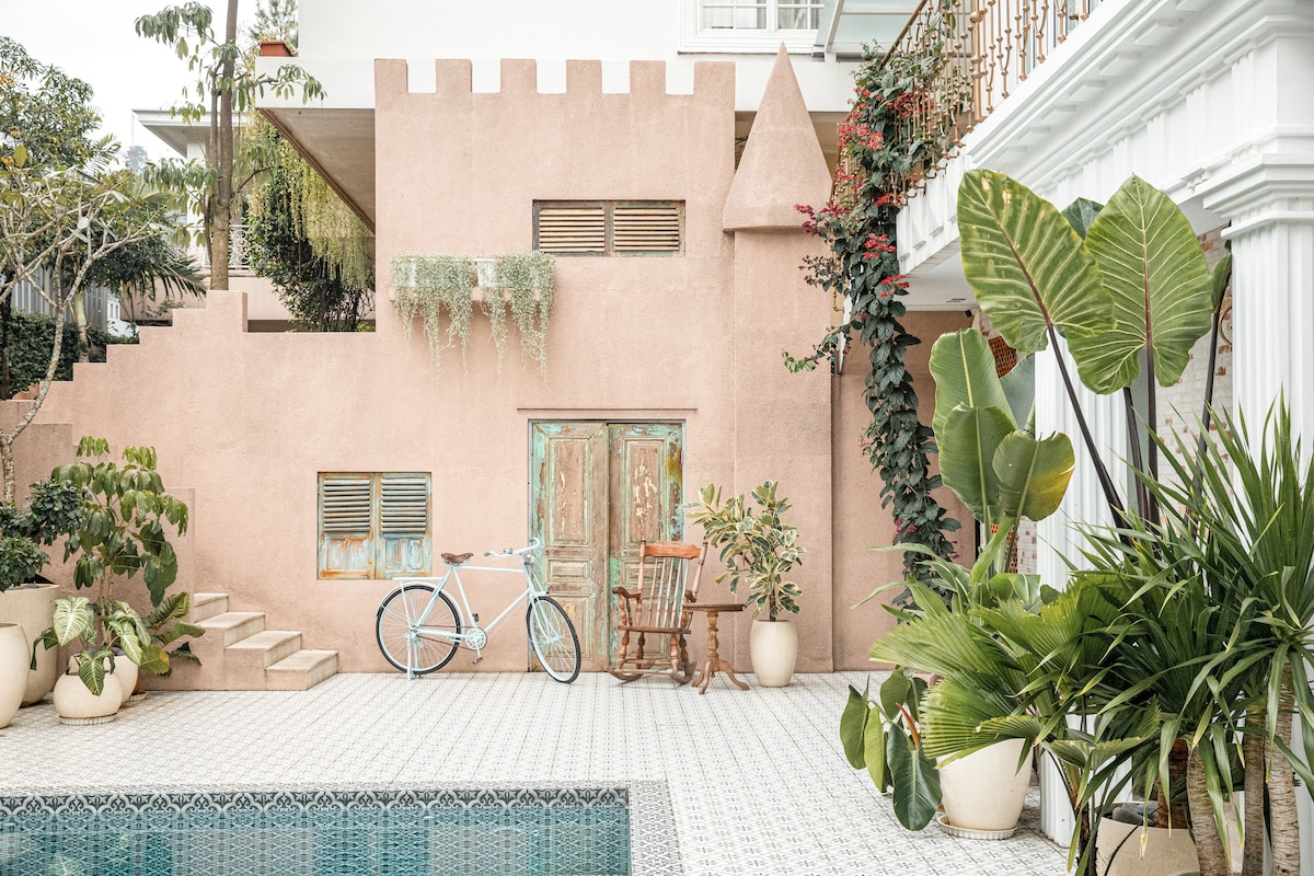 A charming outdoor area features a pink wall designed with castle-like elements. A bicycle rests beside a small wooden table and chair, surrounded by various plants. The light green tiles of the swimming pool reflect the surrounding greenery, enhancing the serene ambiance.