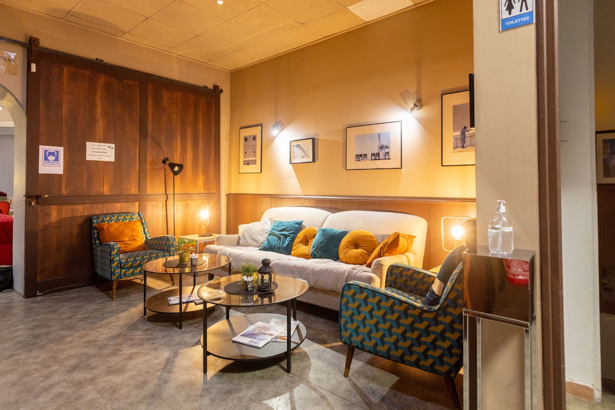 A communal seating area features a white sofa adorned with colorful cushions, surrounded by two patterned armchairs. Coffee tables with elegant designs are positioned in front of the seating. Soft lighting is provided by floor and wall lamps, highlighting the inviting space.