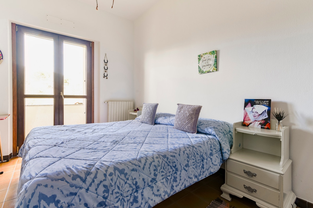 A cozy sleeping area features a spacious bed covered with a light blue quilted blanket. Two decorative pillows are placed at the head of the bed. Natural light is welcomed through the glass double doors that open to a small balcony, creating a bright and airy ambiance.