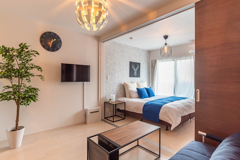 A well-organized living space features a modern television mounted on a white wall. A bed with blue and white bedding is visible in the background, alongside a large window draped with light curtains. A potted plant adds a touch of greenery, complemented by decorative wall accents.