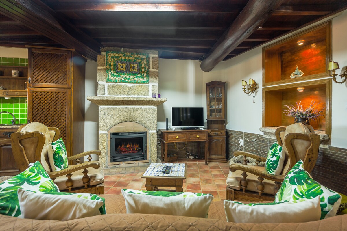 A warm living area is presented with rustic wooden beams overhead and a stone fireplace. Comfortable seating, including patterned chairs and a sofa, surrounds a central wooden coffee table. A flat-screen TV is positioned on an elegant console, integrating modernity within the rustic design.