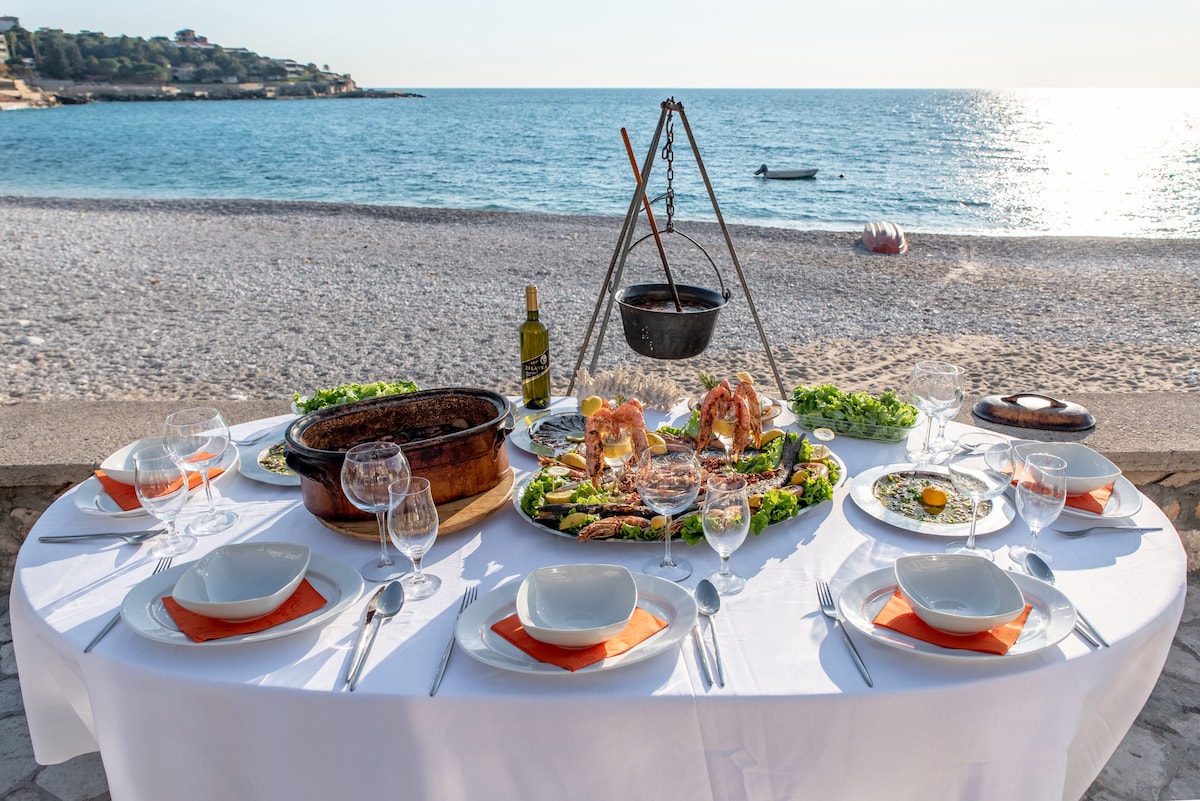 A beautifully arranged outdoor dining table is set with white tableware and orange accents. A variety of seafood dishes are displayed, alongside a bottle of wine. The backdrop features a serene beach and glistening water, enhancing the dining experience.