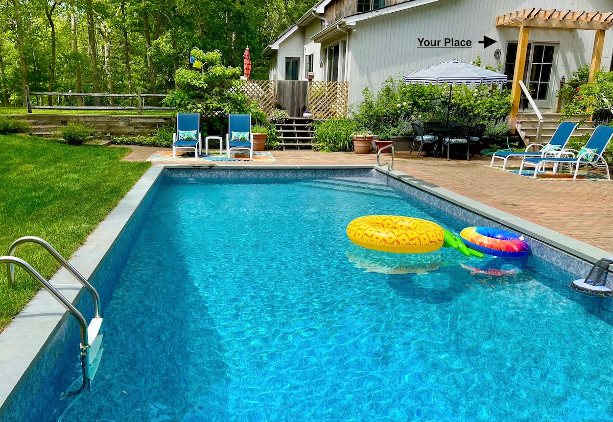 A serene pool area is highlighted by a clear blue swimming pool with colorful floatation devices. Surrounding the pool are lounge chairs and a shaded dining area. Lush greenery creates a private, tranquil atmosphere, enhancing the outdoor retreat experience.
