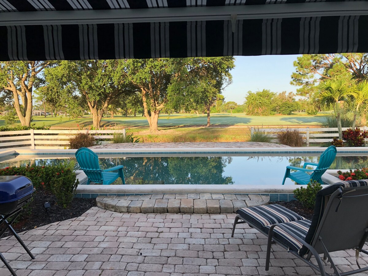 A serene view of a private swimming pool is captured from a covered patio area. Lounge chairs are positioned nearby, surrounded by lush greenery and a golf course in the distance. The pool reflects the clear sky, enhancing the peaceful ambiance.