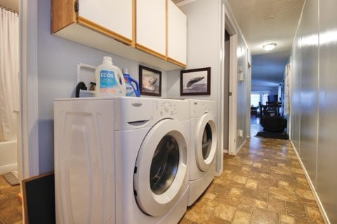 A laundry area is featured along a hallway, showcasing a stacked washer and dryer. Above the appliances, shelves hold cleaning supplies. The tile flooring is visible, leading toward a softly lit living space in the background.