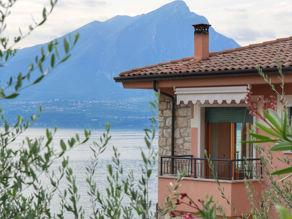 A corner of a beautifully detailed building is visible, featuring a terracotta roof and a balcony with a view of the lake. Lush greenery frames the image, with a backdrop of a mountain range under a cloudy sky.