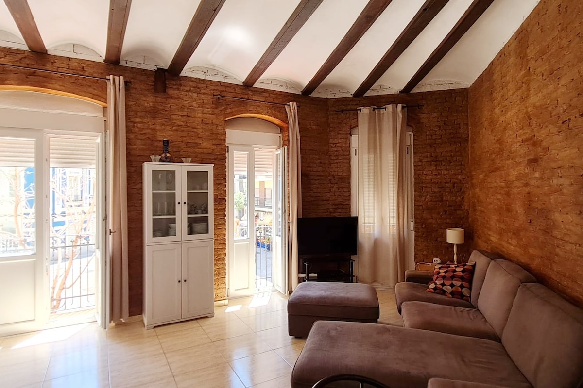 The spacious living area features exposed brick walls and wooden beams. Large windows allow natural light to fill the room, which is furnished with a comfortable sectional sofa and a television. A white cabinet adds storage and visual interest to the space.