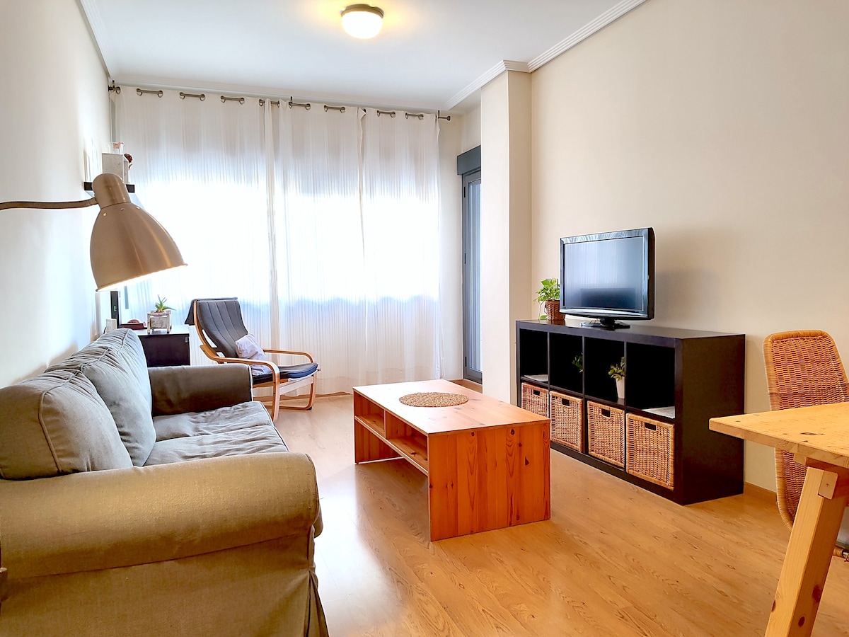 A living area is presented with a comfortable gray sofa and a wooden coffee table at its center. A television stands on a black unit with woven storage baskets. A chair and small table are positioned near a large window, which is framed by sheer white curtains allowing natural light to fill the space.