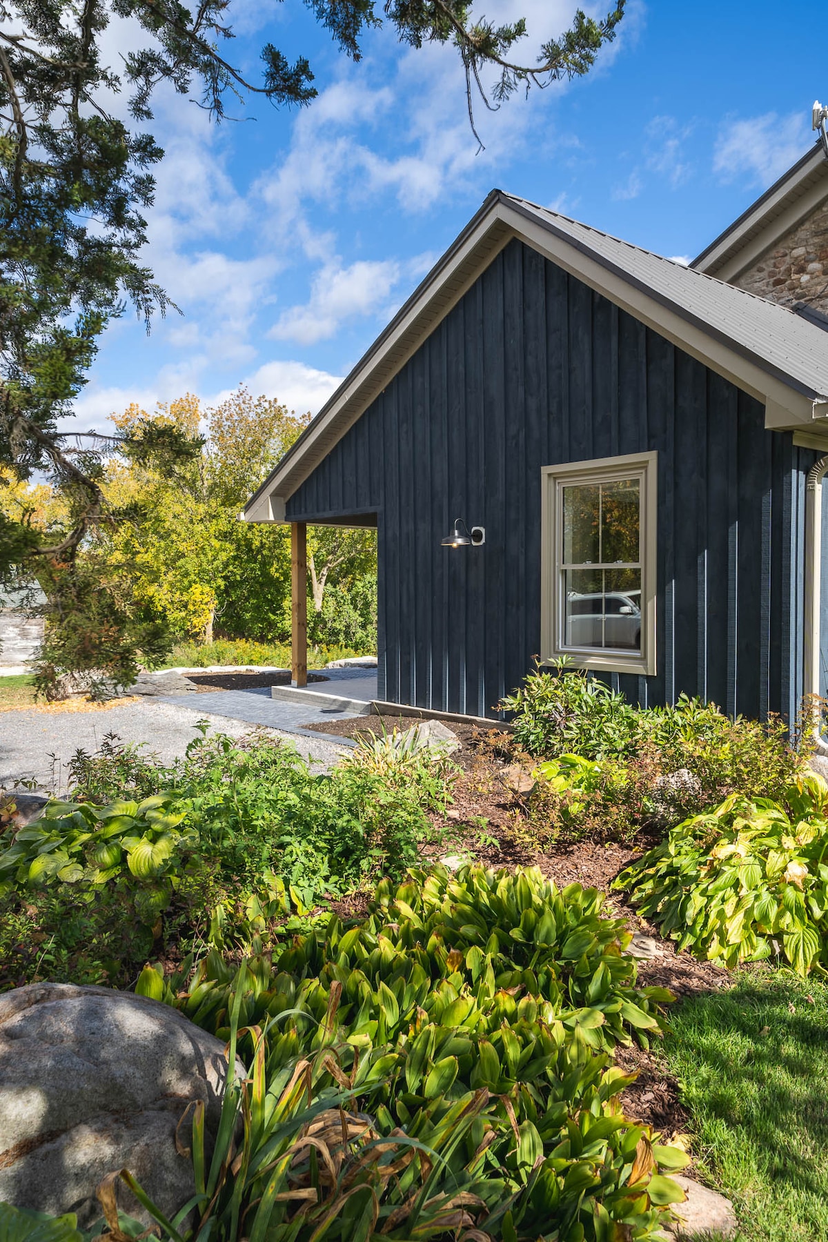 The exterior of the cottage features dark blue siding with a sloped roof and a small overhang. A large window allows natural light to enter, while lush green landscaping and blooming plants surround the home. A pathway leads to a gravel driveway in the background.