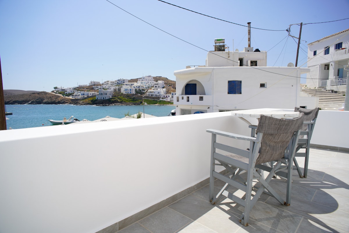 A balcony overlooking the sea features two wooden chairs accompanied by a small table. The view includes whitewashed buildings along the coastline and boats docked in the harbor, set against a clear blue sky.