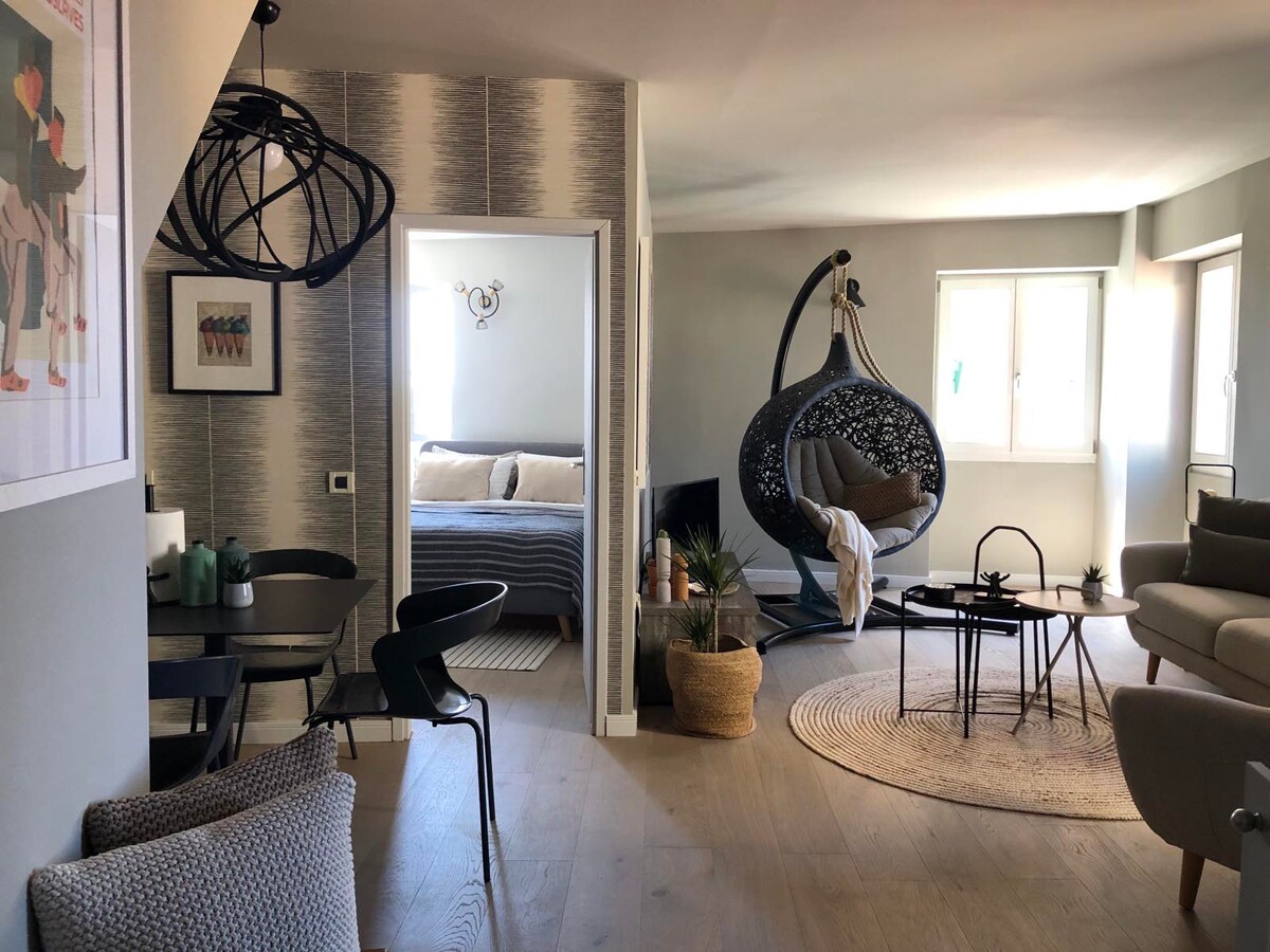 The image captures a modern living space featuring a cozy seating area with a circular rug and a unique hanging chair. A dining table with black chairs is positioned near the entrance, leading to a bedroom area with a spacious bed. Natural light enters through large windows.