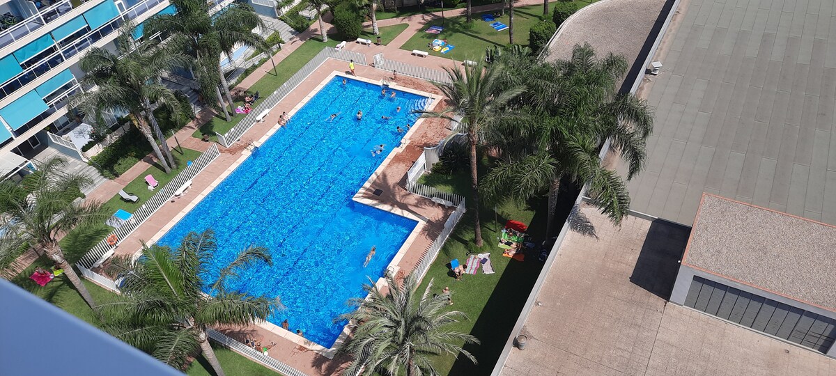 A large outdoor swimming pool is surrounded by neatly manicured lawns and palm trees. Sunbathers relax on colorful lounge chairs along the pool's edge. The clear water is inviting, and a few individuals are swimming in the pool while others enjoy the sun.