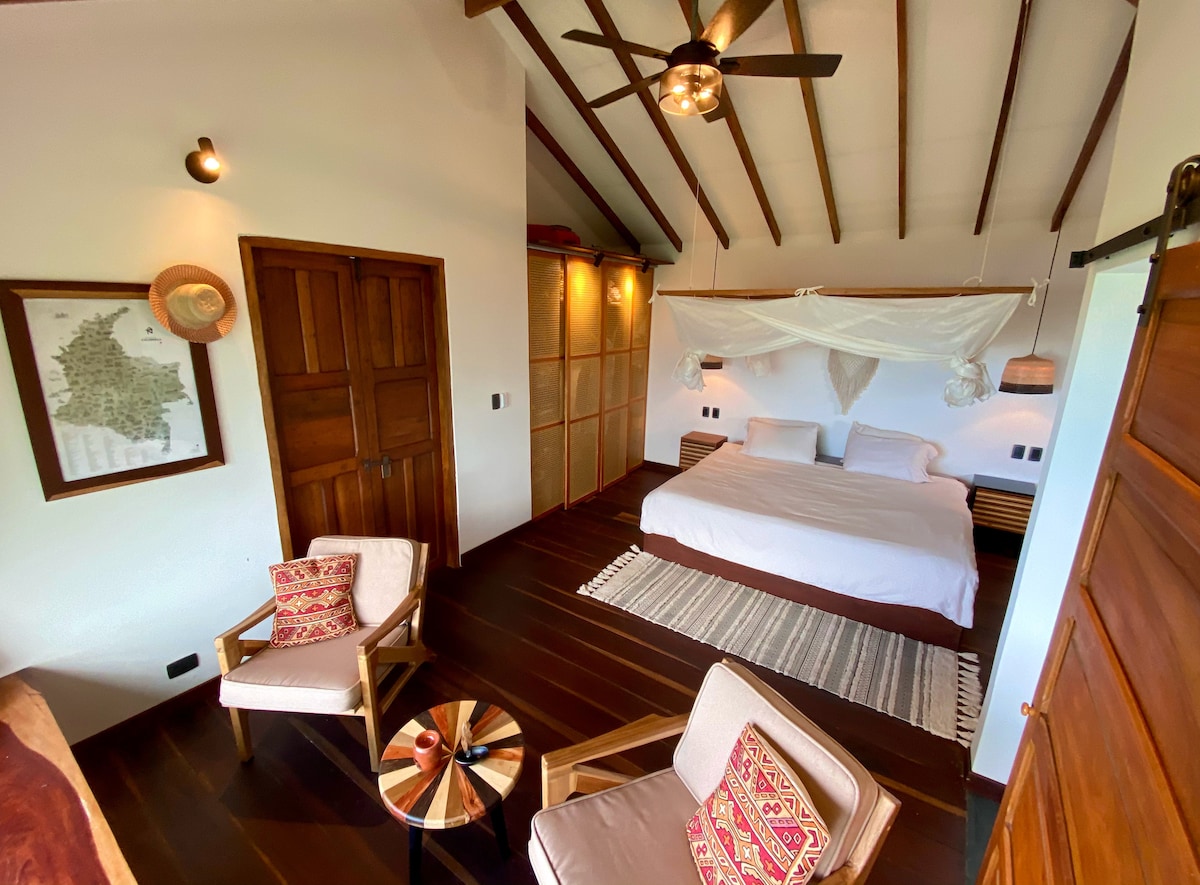 The spacious suite features a king-sized bed draped in a soft canopy. Warm wood tones provide a cozy atmosphere. A comfortable living area is accentuated by two armchairs and a small wooden table. Natural light filters through a window framed by decorative curtains.