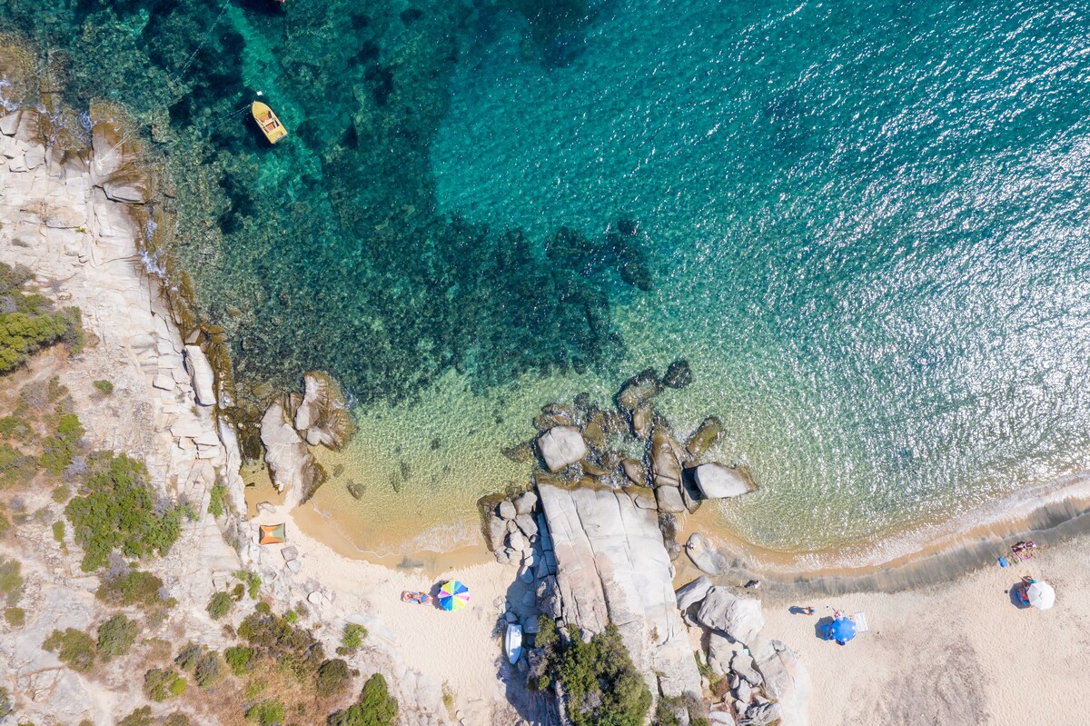 Aerial view of the coastline displaying clear waters transitioning from turquoise to deeper blue. Sandy beach areas are visible, dotted with colorful beach umbrellas. Rocky formations are situated along the shore, providing natural accents to the tranquil landscape.