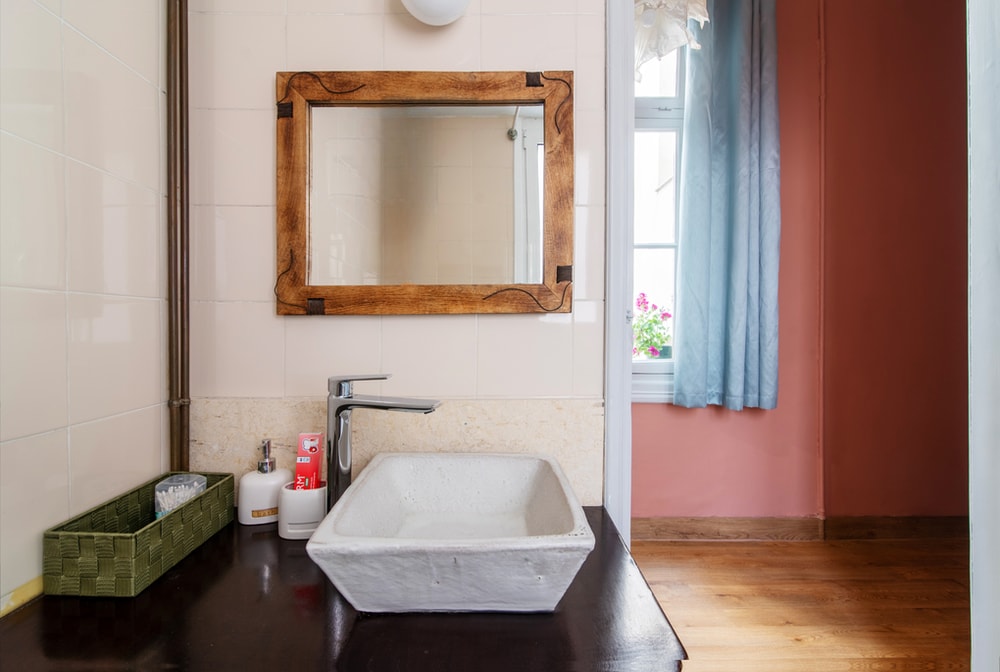 The bathroom features a unique square sink with a textured surface, complemented by a wooden-framed mirror. A small decorative basket holds toiletries, while a bottle of hand soap is also present. Natural light filters through a nearby window adorned with light blue curtains.