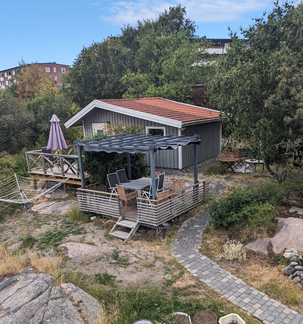 A private terrace is visible in the garden, furnished with a dining table and six chairs, shaded by a pergola. The surrounding greenery adds a natural touch, while a stone path leads toward the cozy cabin featuring a red-tiled roof, creating a welcoming outdoor space.