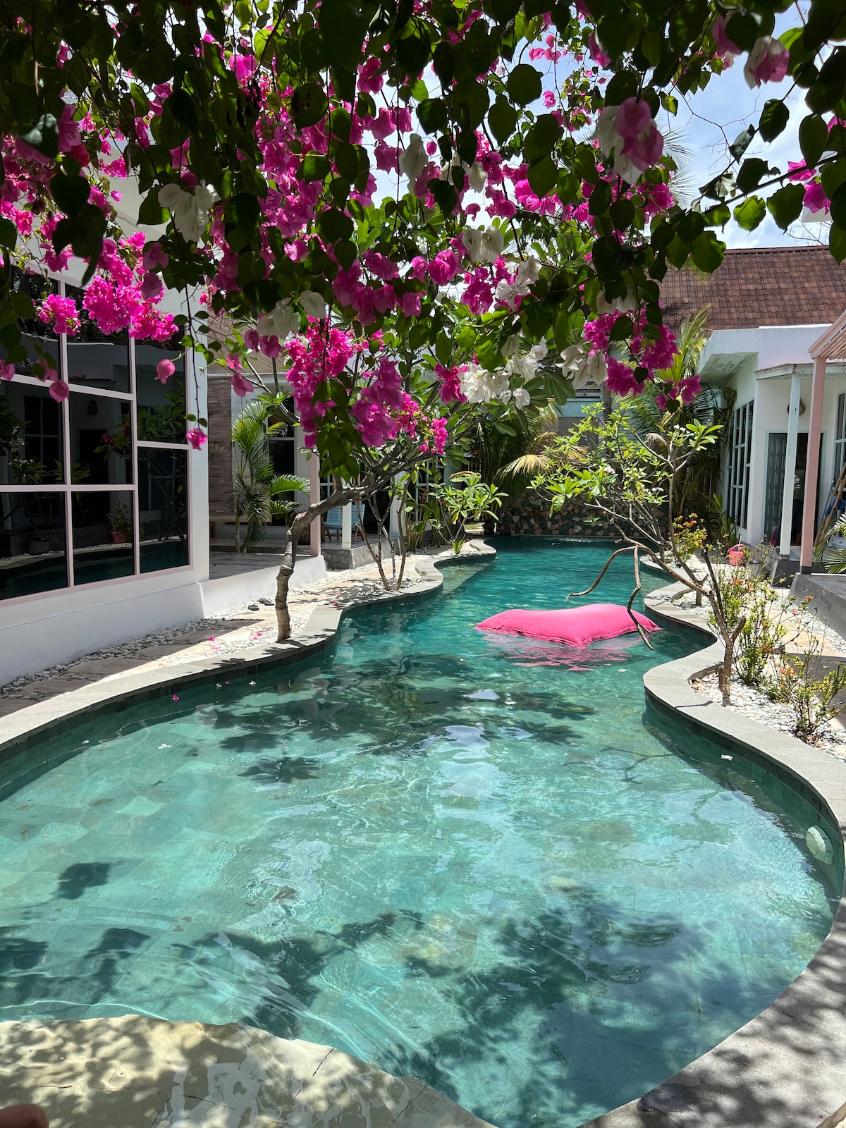 A curvy pool is surrounded by lush greenery and vibrant bougainvillea flowers. The clear water reflects the sunlight, creating a serene atmosphere. Trees and plants are positioned around the pool, enhancing the tropical ambiance of the space.