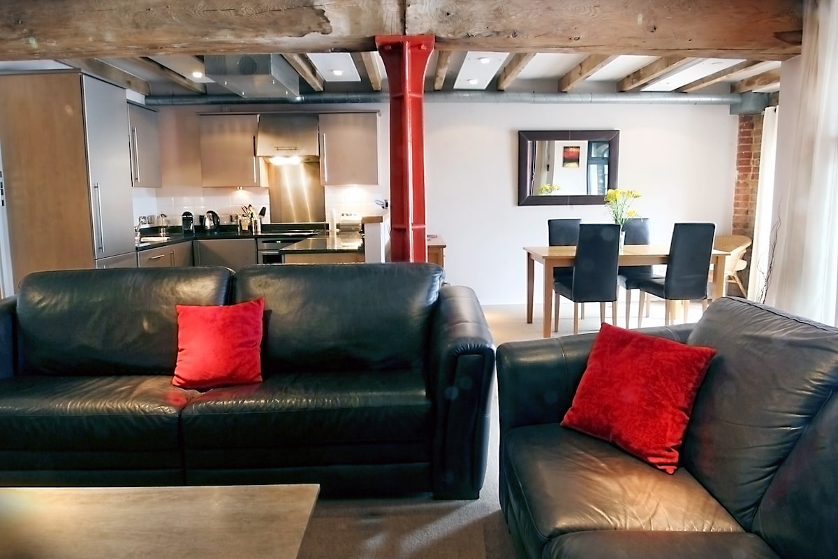 A spacious living area features dark leather sofas adorned with red cushions. A wooden dining table with four black chairs is visible in the background. Exposed wooden beams add character to the open layout, complemented by modern kitchen appliances.